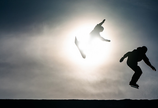 silhouettes of two snowboarders hitting the same jump