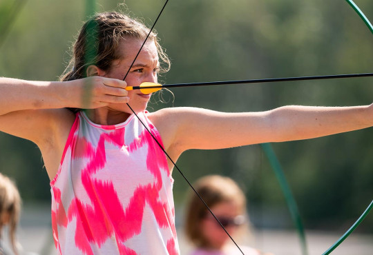 Girl aiming a bow and arrow while at Wilderness summer camp at WinSport